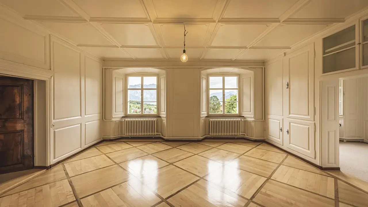 Interior of a room with ornate wooden floor, white paneled walls, two large windows with mountain views, and a single light bulb.