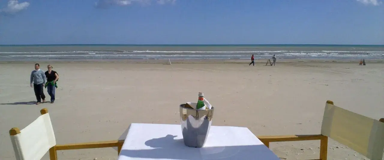 Coppia passeggia sulla spiaggia di Milano Marittima, con un secchiello del ghiaccio e una bottiglia su un tavolo in primo piano.