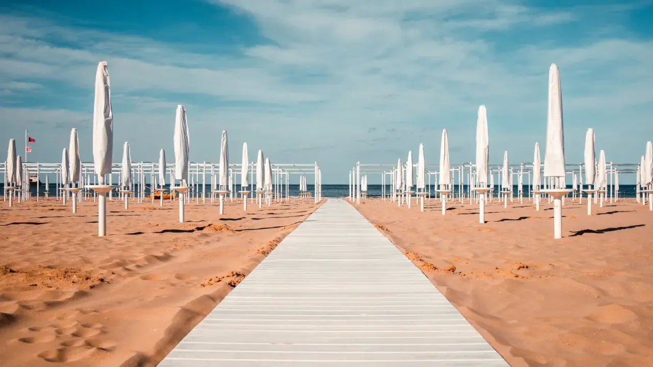 Passerella bianca su sabbia dorata, file di ombrelloni chiusi e strutture bianche sotto un cielo azzurro. Un'immagine serena del mare in Emilia-Romagna.