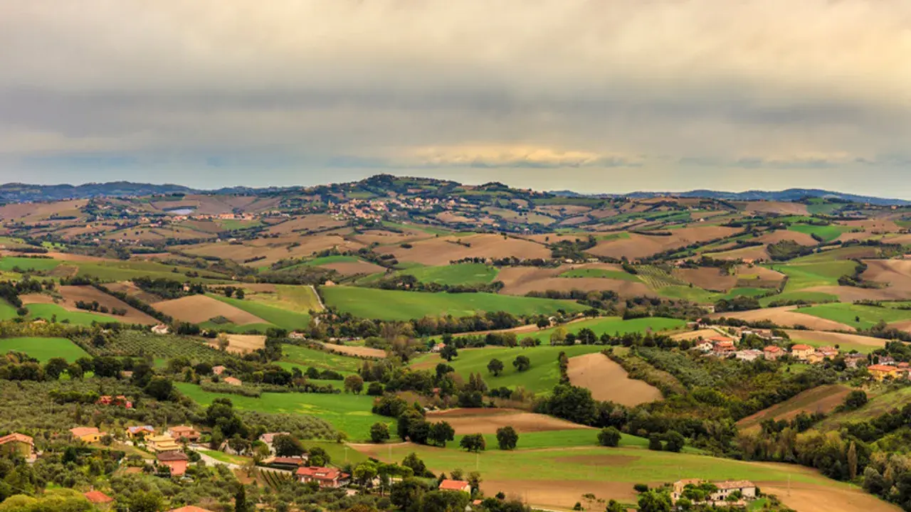 Dolci colline verdi e marroni della Valconca, Rimini, punteggiate da case e alberi, sotto un cielo nuvoloso.