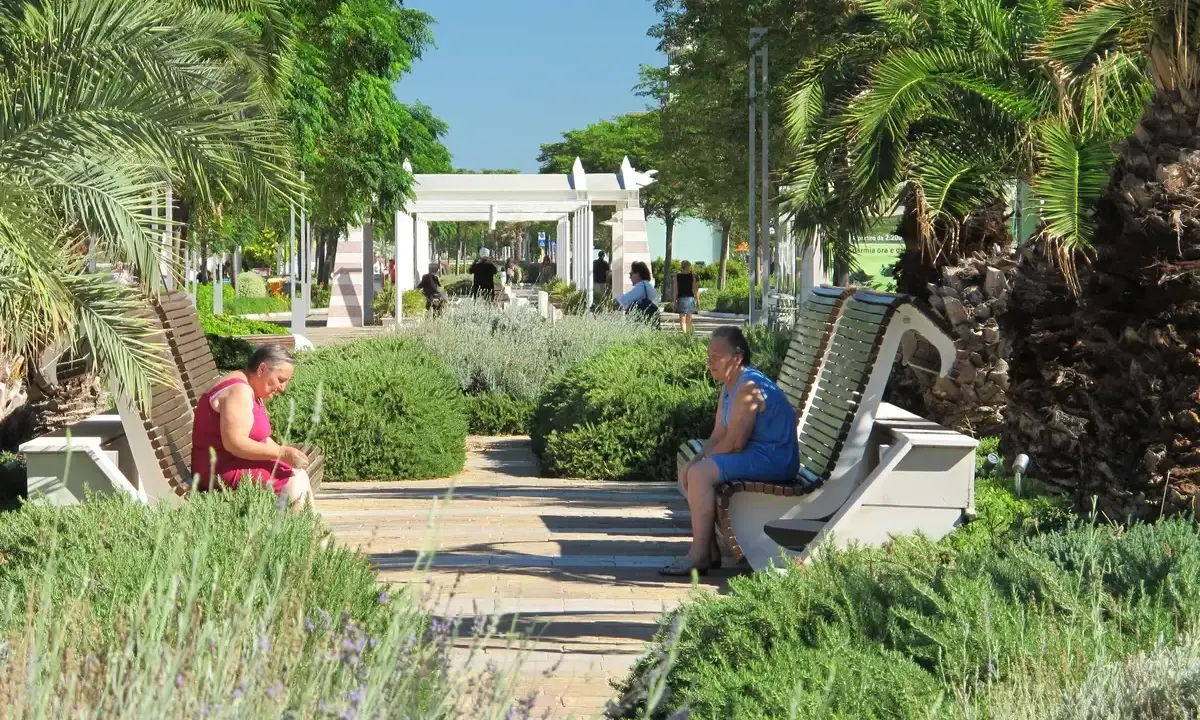 Due donne sedute su panchine in un parco curato, con palme e vegetazione rigogliosa, in una giornata di sole a Riccione centro.