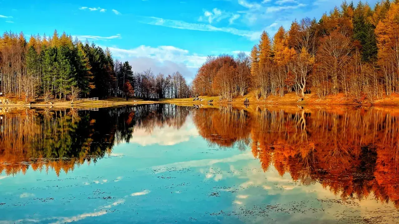 Lago specchiato con alberi autunnali e verdi, ideale per il trekking in Emilia Romagna. Cielo azzurro con nuvole bianche.