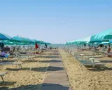 Una passerella di legno conduce alla spiaggia di San Giuliano Mare, fiancheggiata da file di lettini e ombrelloni azzurri sotto un cielo limpido.