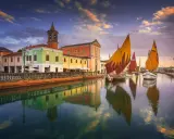 Navi a vela colorate navigano sul canale di Cesenatico al tramonto, con edifici vivaci e riflessi sull'acqua. Meglio Cervia o Cesenatico? Cesenatico offre questo spettacolo.
