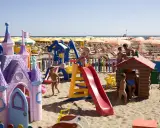 Bambini giocano in un'area giochi colorata sulla sabbia, con castello e scivoli, mentre ombrelloni gialli e lettini creano un'atmosfera da **sunday beach Rimini**.