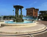 Fontana con statue di donne e tartarughe in piazza Primo Maggio Cattolica, con un edificio in mattoni sullo sfondo.