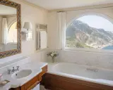 Un bagno di lusso con vasca, lavandino e specchio decorato, che offre una vista mozzafiato sulle montagne e sul mare. Tra i bagni più belli ed eleganti.