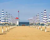 Filetto di ombrelloni a righe blu e bianche sulla spiaggia di Rivabella di Rimini, con mare calmo e cielo azzurro.