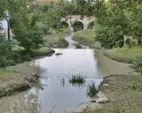 Un fiume serpeggia verso un antico ponte ad archi, evocando il **passaggio del Rubicone**. Vegetazione lussureggiante e acque tranquille.