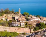 Borgo antico con campanile affacciato sul mare Adriatico. Il centro storico del lungomare Gabicce Mare, con tetti rossi e vegetazione rigogliosa, si estende verso l'azzurro intenso dell'acqua.