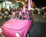 Un'auto rosa shocking sfila a Rimini durante la Notte Rosa, con un pupazzo a forma di pantera rosa sul tetto e un uomo al volante vestito a tema.