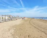 Spiaggia di Ravenna Lido Adriano con file di ombrelloni e lettini, bagnanti che passeggiano sul bagnasciuga sotto un cielo azzurro.