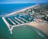 La darsena di Rimini, con le sue barche ormeggiate, si estende lungo la costa adriatica, tra la spiaggia affollata e il mare azzurro.