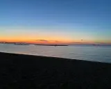 Tramonto sulla spiaggia di Rimini, con il cielo che sfuma dall'arancione al blu. Due persone passeggiano sulla sabbia scura, mentre in mare si vedono barche a vela.