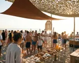 Un gruppo di persone festeggia su una spiaggia di Rimini, con un buffet di dolci e caffè. Alcuni fanno yoga, altri si godono il sole.