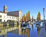 Traditional sailboats with colorful sails line a canal in Cesenatico, Italy, with buildings and a church reflected in the water.