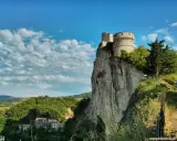 La fortezza di San Leo domina un picco roccioso, con un borgo sottostante e un cielo azzurro punteggiato di nuvole.
