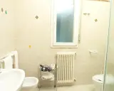 Bagno 79 Rimini: lavabo, bidet, WC, scaldasalviette e finestra. Asciugacapelli e rotolo di carta igienica su carrello.