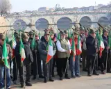 Uomini con cappelli alpini e bandiere italiane partecipano all'adunata alpini a Rimini, con un ponte storico sullo sfondo.