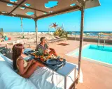 Coppia rilassata su lettini con vassoio di frutta e caffè, con piscina e spiaggia di Rimini sullo sfondo.