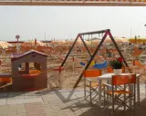 Area giochi e lettini sulla spiaggia 106 di Riccione, con ombrelloni arancioni e bianchi, altalena e casetta per bambini.