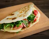 Piadina romagnola, un classico dal sapore autentico, ripiena di prosciutto, formaggio, rucola e pomodorini.