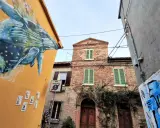 Un murale di balena colorata decora un edificio giallo a San Giuliano a Mare. Vicino, un edificio in mattoni con persiane verdi e un cartello di area pedonale.