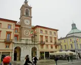 La piazza Rimini, con il suo imponente campanile e gli edifici storici, accoglie visitatori e residenti sotto un cielo nuvoloso.
