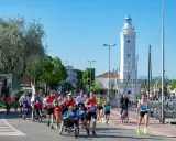 Atleti corrono lungo il percorso della Rimini Marathon, con il faro bianco e il porto sullo sfondo.