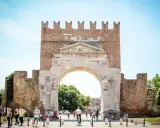 L'imponente arco d'augusto rimini, con le sue antiche pietre e la torre in mattoni, fa da sfondo a una vivace piazza con persone che passeggiano e ciclisti.