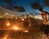 Spettacolo di fuoco sulla spiaggia di Rimini, estate. Artista si esibisce tra torce e fuochi d'artificio, con pubblico e luci in lontananza.