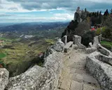 Sentiero in pietra conduce al **San Marino castle**, con vista panoramica sulla valle verdeggiante e colline ondulate sotto un cielo nuvoloso.