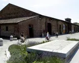Passeggiata in bicicletta davanti ai Magazzini del Sale di Cervia, con persone che pedalano su un lungomare acciottolato, tra aiuole fiorite e antichi edifici in mattoni.