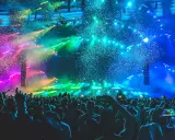 Folla in delirio a un concerto a Rimini, con coriandoli colorati e luci psichedeliche che illuminano la scena.