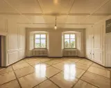 Interior of a room with ornate wooden floor, white paneled walls, two large windows with mountain views, and a single light bulb.
