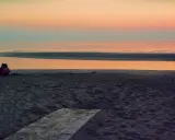 Alba a Rimini: una coppia seduta sulla sabbia guarda il mare calmo e il cielo colorato.