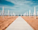 Passerella bianca su sabbia dorata, file di ombrelloni chiusi e strutture bianche sotto un cielo azzurro. Un'immagine serena del mare in Emilia-Romagna.