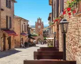 Un vicolo acciottolato a Gradara, con edifici in mattoni, fiori rossi e un campanile con orologio. Perfetto per scoprire cosa vedere.
