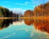 Lago specchiato con alberi autunnali e verdi, ideale per il trekking in Emilia Romagna. Cielo azzurro con nuvole bianche.
