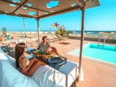 Coppia rilassata su lettini con vassoio di frutta e caffè, con piscina e spiaggia di Rimini sullo sfondo.