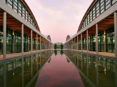 Ingresso moderno della fiera a Rimini, con un lungo specchio d'acqua che riflette le arcate e il cielo rosa del tramonto.