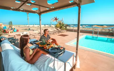 Coppia rilassata su lettini con vassoio di frutta e caffè, con piscina e spiaggia di Rimini sullo sfondo.