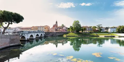 Ponte di Tiberio e il fiume Marecchia a Rimini a settembre, con riflessi sull'acqua e un cielo azzurro con nuvole bianche.