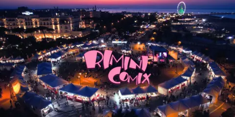 Un vivace evento serale a Rimini Comix, con bancarelle illuminate, una ruota panoramica e il logo rosa che domina la scena.