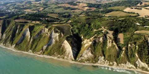 Scogliere a picco sul mare turchese a Gabicce Mare, Marche. Campi coltivati e vegetazione rigogliosa adornano le colline retrostanti.