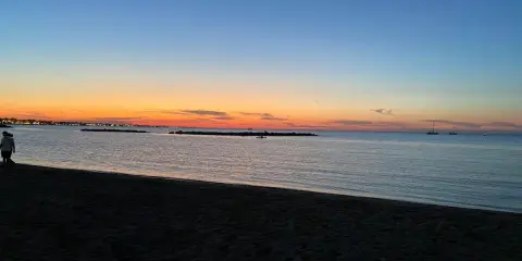 Tramonto sulla spiaggia di Rimini, con il cielo che sfuma dall'arancione al blu. Due persone passeggiano sulla sabbia scura, mentre in mare si vedono barche a vela.