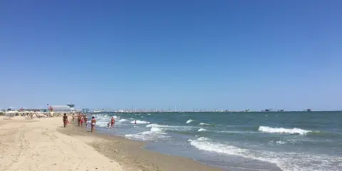 Spiaggia affollata a Ravenna, con gente che passeggia sulla sabbia e nel mare. All'orizzonte, barche e un cielo azzurro intenso.