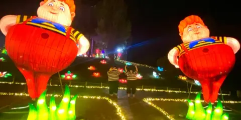 Gigantesche lanterne di bambini paffuti illuminano un sentiero a Wonderland Rimini, con due persone che salutano tra fiori luminosi.