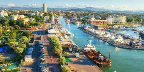 Vista aerea del porto di Rimini con barche ormeggiate, edifici e montagne sullo sfondo.