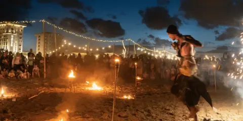 Spettacolo di fuoco sulla spiaggia di Rimini, estate. Artista si esibisce tra torce e fuochi d'artificio, con pubblico e luci in lontananza.